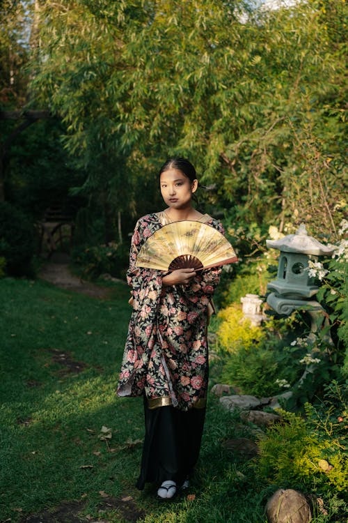 a young asian girl stands in a lush green garden, elegantly dressed in a floral kimono and holding a hand fan. she gazes at the camera with a serene expression, creating a peaceful and traditional scene. the setting enhances the image's cultural and aesthetic qualities.