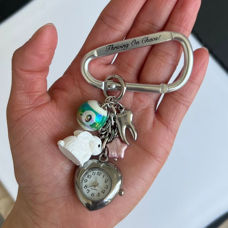 a person is holding a silver carabiner keychain with various charms attached in their hand. the charms include a heartshaped watch, a 9ball, a tooth, a rabbit, and a star. the carabiner has the words "thriving on chaos!" engraved on it.