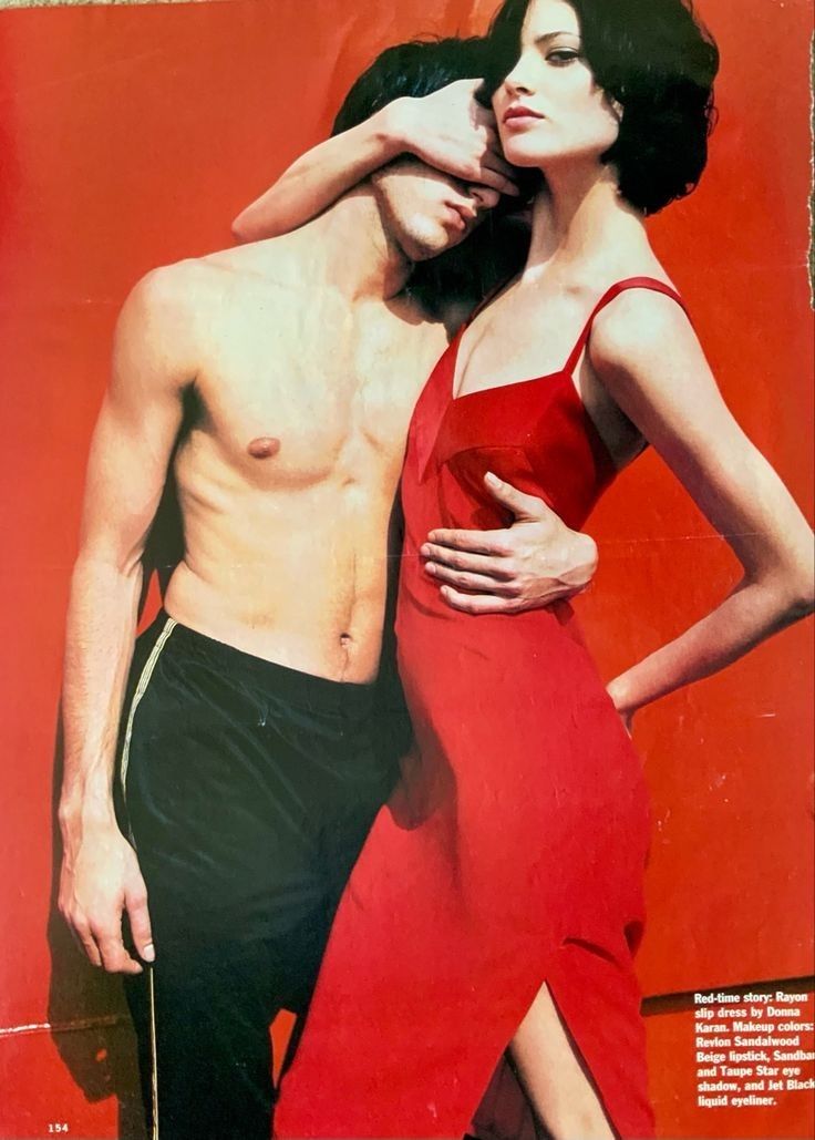 a vintage fashion editorial featuring a woman in a red rayon slip dress by donna karan, with the makeup details of revlon sandalwood beige lipstick, sandbo and taupe star eye shadow, and jet black liquid eyeliner. she is standing next to a shirtless man who has his eyes covered by her hand, and she embraces him. the background is red, and the image appears to be from a 1990s magazine spread.