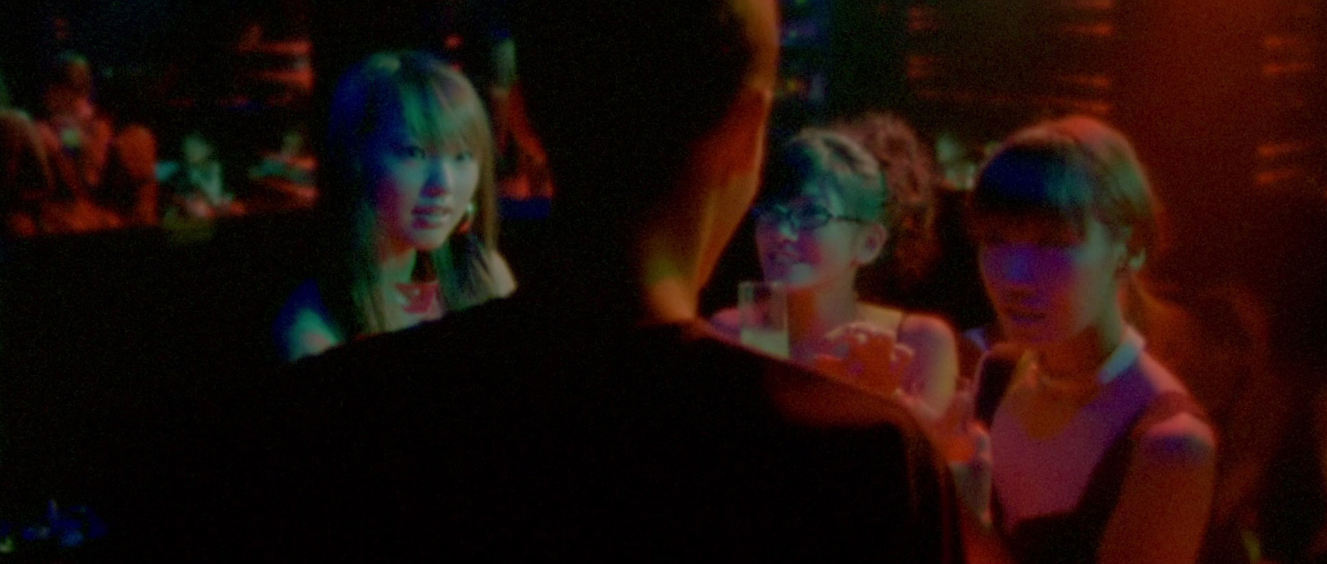 the image shows a group of young people in a dimly lit nightclub. the perspective is from over the shoulder of a person in the center, facing away from the camera. three young women stand in front of them, illuminated by colorful lights. the atmosphere suggests a lively and social setting.