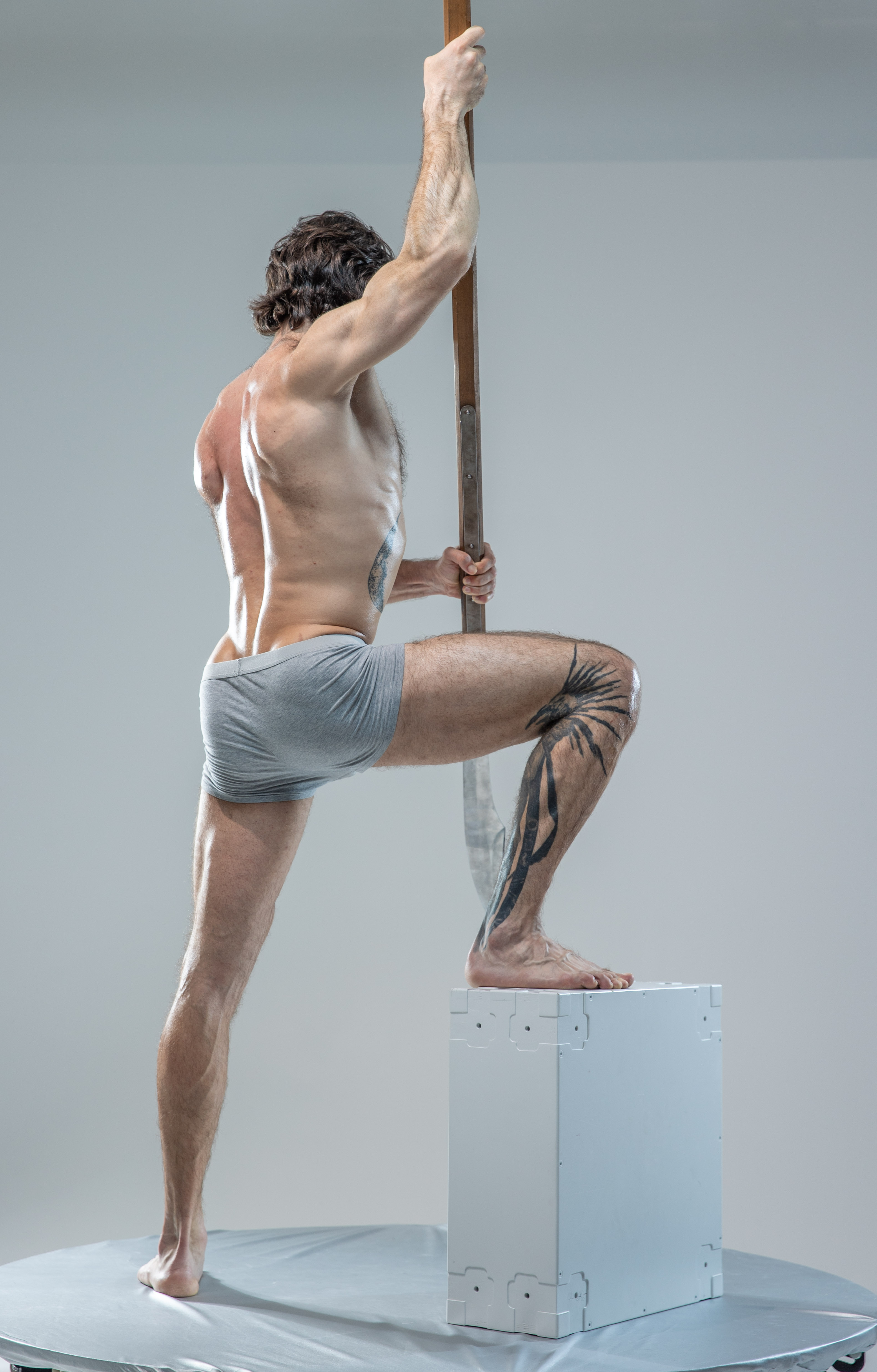 a muscular man with tattoos is standing in a studio setting, his back to the camera. he is wearing grey underwear and holding a long, wooden staff. he is posing with one foot on a white cube, his body creating a dynamic line.  the lighting is soft and diffused, highlighting his musculature.  the image appears to be a reference for artists.