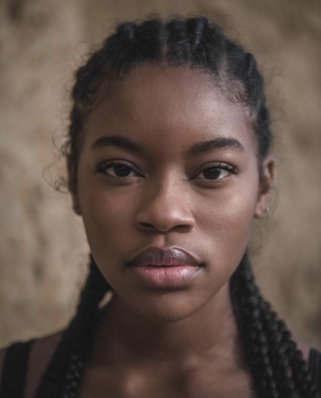 a closeup portrait of a young black woman with braided hair. she has dark skin, a serious expression, and is facing the camera directly. the background is a neutral color, and the lighting appears natural.