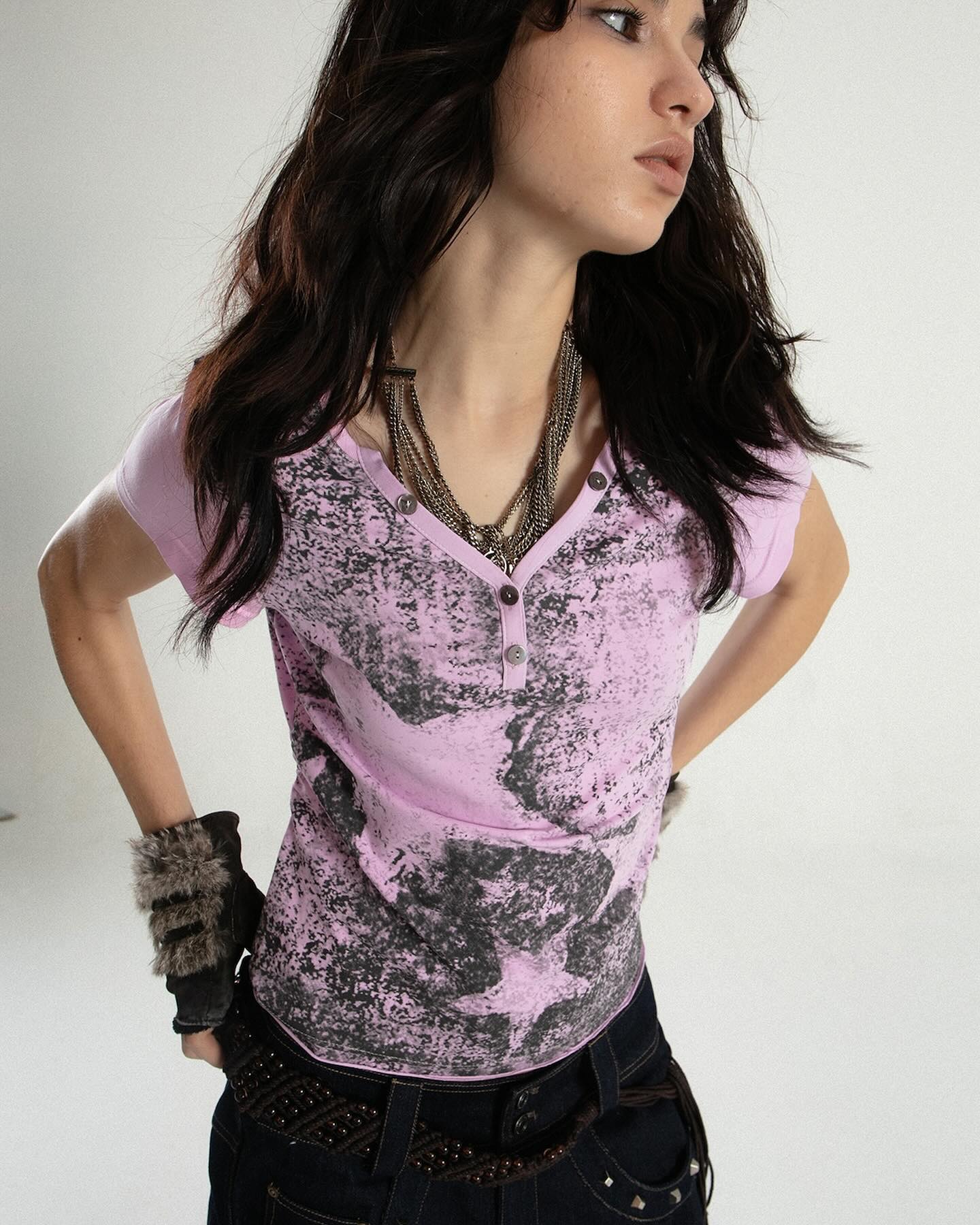closeup shot of a young woman with long dark hair wearing a pale lavender henleystyle tshirt with a dark graphic print. she is also wearing dark wash jeans, a beaded belt, and several necklaces and bracelets.  she has on furtrimmed gloves and is posing against a white background. the image is welllit and shows off the details of her clothing and accessories.