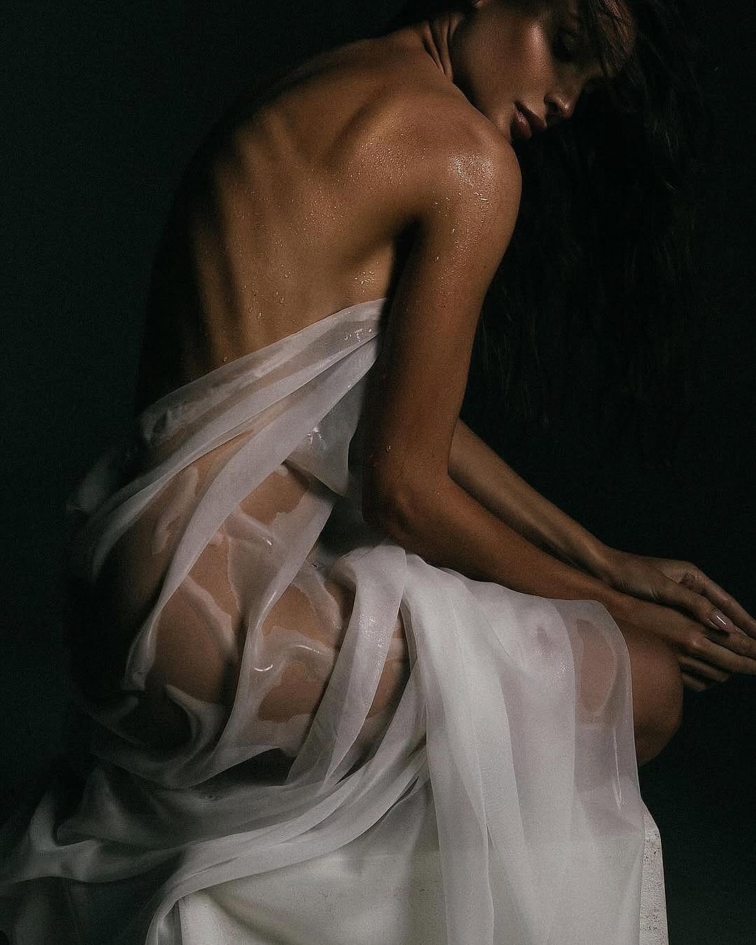 the image is a closeup, artistic portrait of a woman with dark hair and brown skin. she is partially draped in a sheer, white fabric that clings to her wet body, showcasing the water droplets on her skin. the background is dark and shadowy, creating a contrast with the lighter tones of the woman and the fabric.  the overall mood is sensual, mysterious, and elegant. the pose and lighting suggest a fashion or fine art photography context.