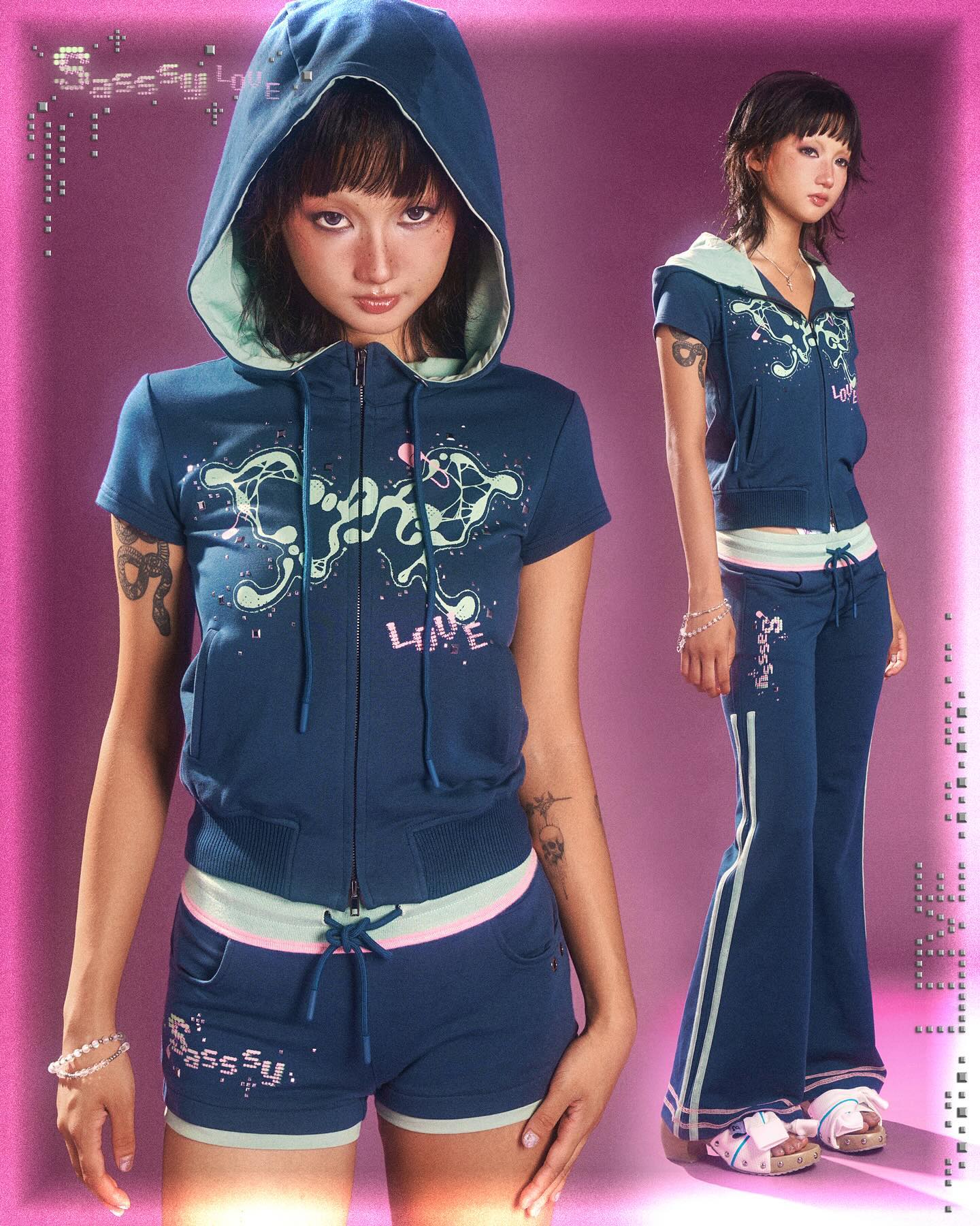 the image showcases two models wearing a teal and pastelcolored tracksuit.  the tracksuit features a shortsleeved zipup hoodie with a graphic design and matching shorts. one model is wearing the full tracksuit, while the other is shown in the hoodie and shorts. the overall style is reminiscent of y2k or early 2000s fashion. the background is a bright, pinkpurple gradient. both models have dark hair,  and one model has visible tattoos on her arms. the image has an overlay of text ("sassy luv") that is styled similarly to the graphic design on the clothing.  the overall feeling is playful and stylish, highlighting the clothing's unique design.