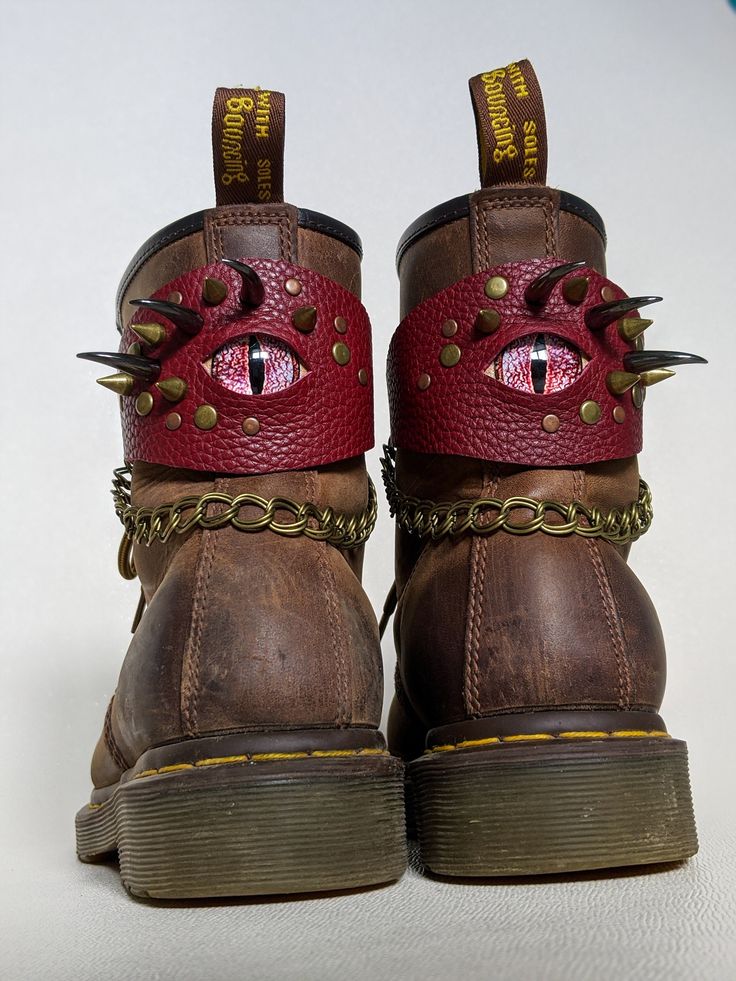 a pair of brown leather dr. martens boots with dragon eye and scale details. the boots have a red dragon eye design on the ankle with metal spikes and a gold chain. the boots are sitting on a white background.