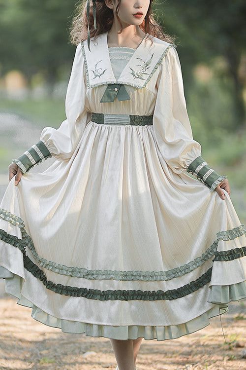 a woman in a vintageinspired dress stands outdoors. the dress is a light cream color with green accents, featuring a sailor collar, long sleeves, and a ruffled skirt. the dress falls to her ankles, and the skirt has multiple layers of ruffles. she is posing with her arms outstretched, showing off the dress's flowy silhouette. the overall aesthetic of the image is romantic and whimsical, with a focus on nature and vintage fashion.