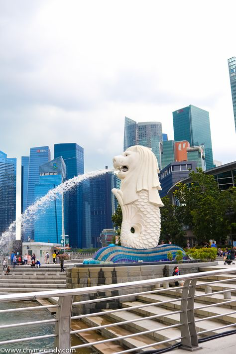 the image showcases the iconic merlion statue in singapore, a halflion, halffish creature spouting water from its mouth. the statue is prominently featured in the foreground, with a vibrant fountain at its base.  behind the merlion, a stunning backdrop of modern, highrise skyscrapers, characteristic of singapore's city skyline, is visible.  people can be seen in the background, relaxing in the parklike area surrounding the merlion and fountain. the overall ambiance is bright and sunny, suggesting a typical day in singapore. a sleek, metallic railing and steps are in the foreground, adding to the contemporary feel of the scene.
