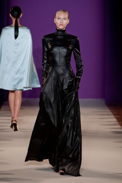 a runway shot featuring two models. the focus is on a woman in a floorlength black leather dress with a high turtleneck.  the dress is sleek and formfitting.  she has a sophisticated and elegant demeanor. in the background, slightly out of focus, is another woman wearing a light blue cape. the runway is lightcolored, and the backdrop is a deep purple. the overall atmosphere is one of high fashion.