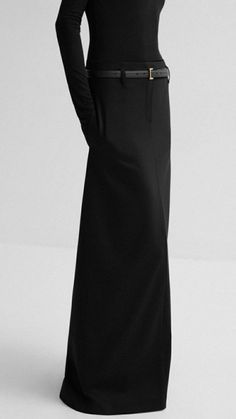 a woman poses in a black longsleeved top and long, black skirt.  she is wearing a black leather belt with a gold buckle and is standing in a studio against a white background.