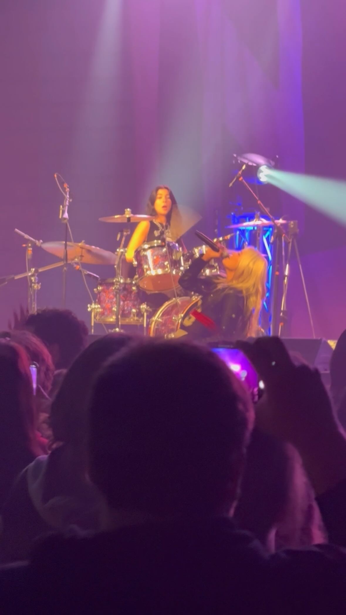 a live concert is taking place with a female drummer and a singer performing on stage. the drummer is seated behind her kit, and the singer is positioned in front, engaging with the audience. the stage is illuminated with vibrant purple lighting and a spotlight. a large crowd is present, with some individuals holding up their phones to capture the moment.