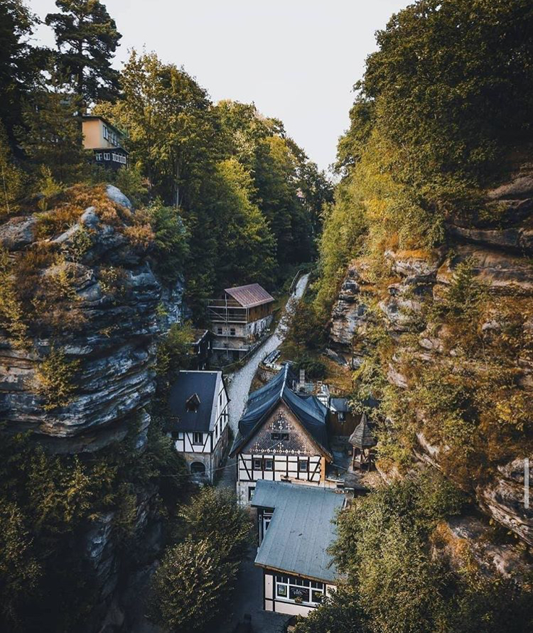 an aerial view of a village nestled between rock formations and trees. the houses feature halftimbered architecture and are surrounded by lush greenery. a narrow road winds through the village. the surrounding landscape includes mountains and a dense forest.