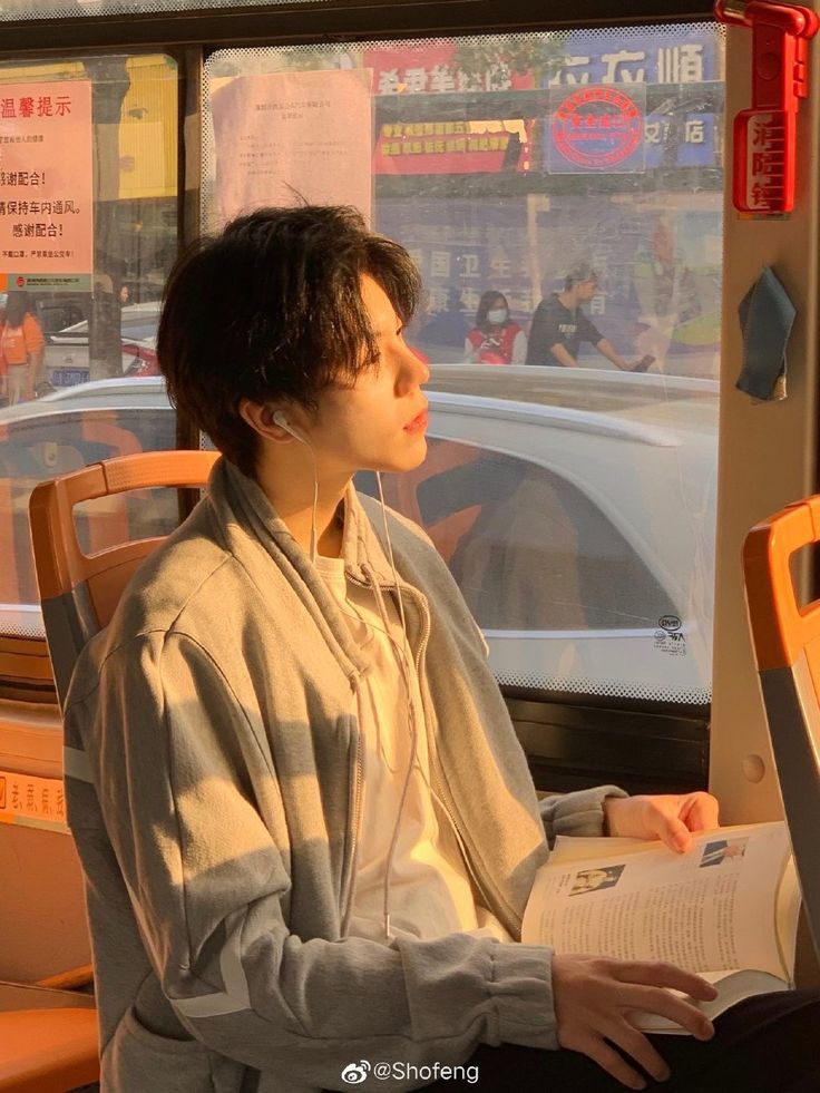 young man with short, dark hair is seated on a bus, engrossed in a book bathed in warm, golden sunlight streaming through the window. he's wearing a light blue and white jacket and has white earphones in. the setting sun casts a soft glow on his face, highlighting a contemplative expression as he reads. outside the window, a bustling city street scene unfolds with blurred figures and vehicles passing by.