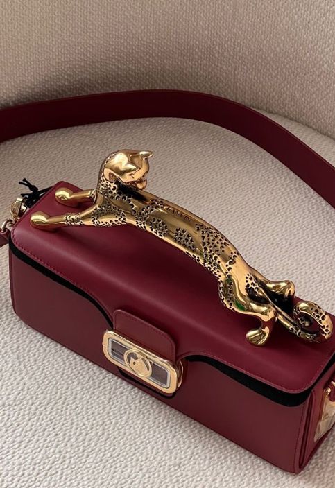 a maroon leather handbag with a gold leopard motif handle and shoulder strap is displayed on a neutral textured surface. the bag has a clasp closure with gold detailing. the leopard handle is sculpted and detailed with spots. the shoulder strap matches the bag's color.