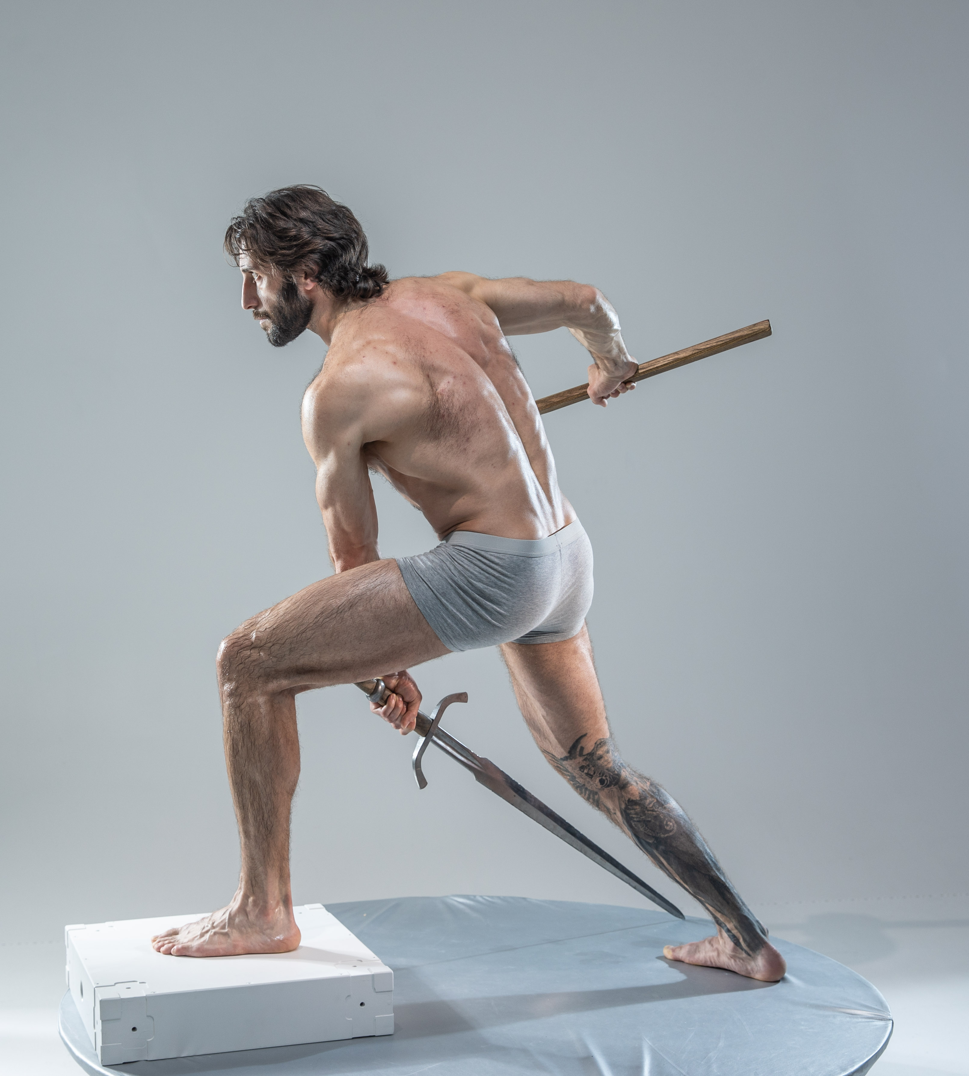 a muscular man with long hair and tattoos stands barefoot on a white platform in a studio setting. he adopts a dynamic pose, his body angled as if preparing to strike.  he holds a short sword in his right hand, the tip resting on the platform, and a wooden staff in his left hand, held high.  he wears only grey underwear, his physique illuminated by soft studio lighting. the background is a plain light grey.  the overall image evokes themes of power, action, and masculinity.