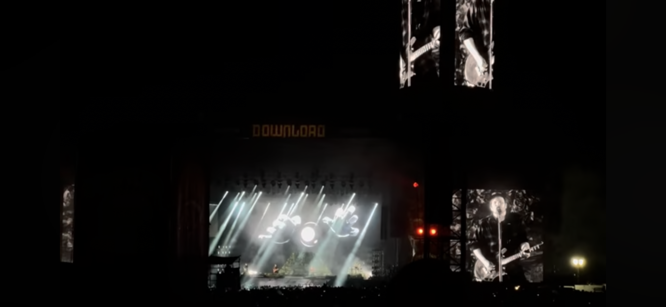 a nighttime shot of a rock concert stage with the name "download" visible at the top. the stage is illuminated by bright lights and there is a large crowd in front of it.  there are also large video screens on either side of the stage, displaying a closeup of a musician playing guitar. the scene is dark, but the lights and screens make it clear that this is a major rock concert event.