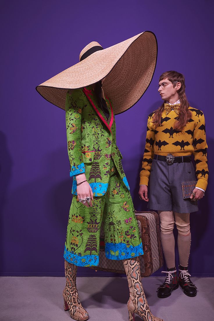 two models stand against a purple backdrop, showcasing gucci's collection. a woman wears a vibrant green suit with a striking floral pattern, accented with turquoise trim. her outfit is completed with an oversized straw hat and eyecatching snakeskin boots. the male model beside her sports a yellow batpatterned sweater and grey shorts, exuding a contrasting yet equally stylish look. a vintagestyle suitcase adds to the scene’s overall aesthetic. the image beautifully captures the collection's blend of bold patterns and unique silhouettes.