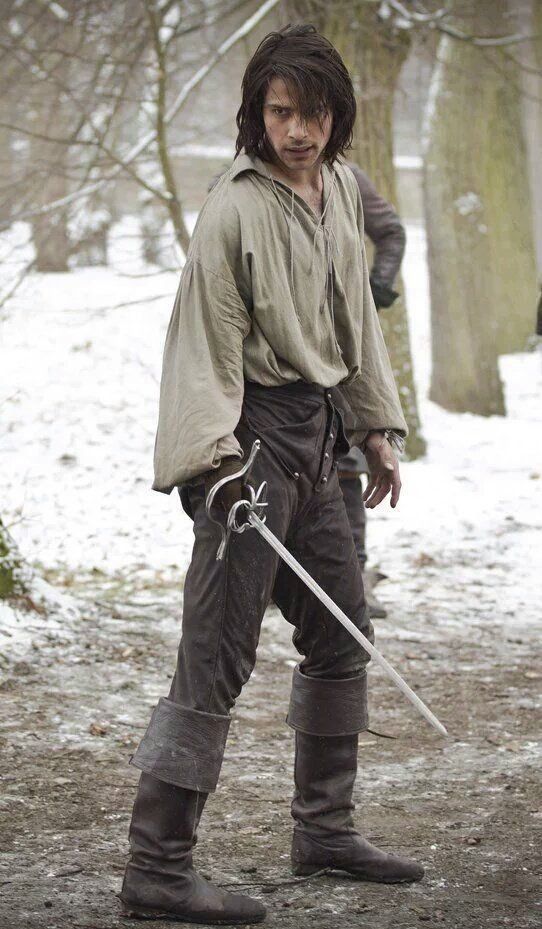 a man in a historical costume, possibly a musketeer, stands in a snowy forest. he holds a sword in his right hand, and is looking directly at the camera. the lighting is soft and overcast, giving the image a cold and dramatic feel.