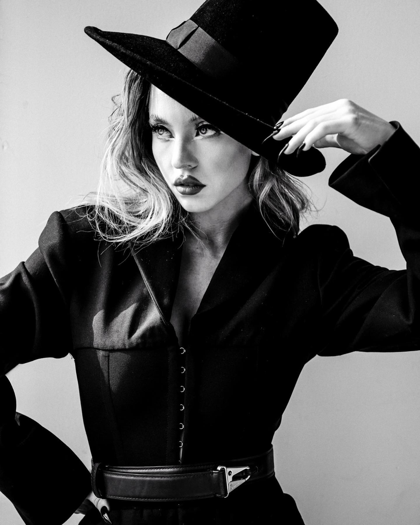 the image is a black and white, closeup portrait of a fashionable woman. she is wearing a tall black top hat, a longsleeved black jacket with a corsetlike bodice, and a black leather belt.  her hair is styled in loose waves, falling around her shoulders. she looks directly at the camera with a somewhat serious and intense expression.  the lighting is dramatic and focused on the model, creating contrast and depth. the overall style is high fashion and evokes a sense of elegance and mystery. the background is a plain, lightcolored wall, keeping the focus solely on the model and her attire.