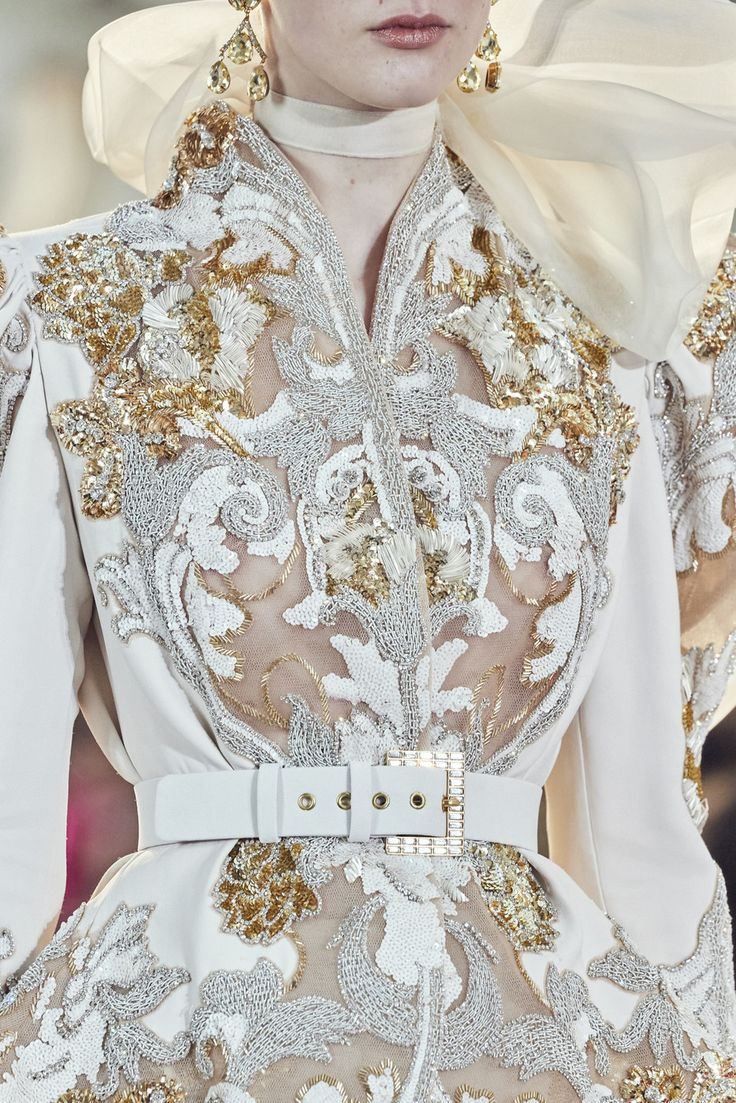 the image showcases a closeup detail of a stunning haute couture gown. the dress is predominantly white, featuring intricate gold and white beading and embroidery that creates a baroqueinspired floral pattern. the embroidery is densely layered, adding significant texture and opulence to the garment.  a crisp white belt cinches the waist. the model wears large, ornate gold earrings. the fabric appears to be a combination of sheer and opaque layers, allowing the intricate work to be fully visible. the overall style is extremely elegant and luxurious, suggesting a highend designer piece likely shown on a runway.