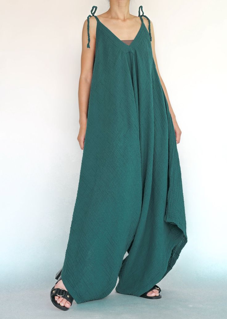 a woman wearing a teal, flowy, linen jumpsuit with thin straps, standing against a white background.  she is wearing black sandals.