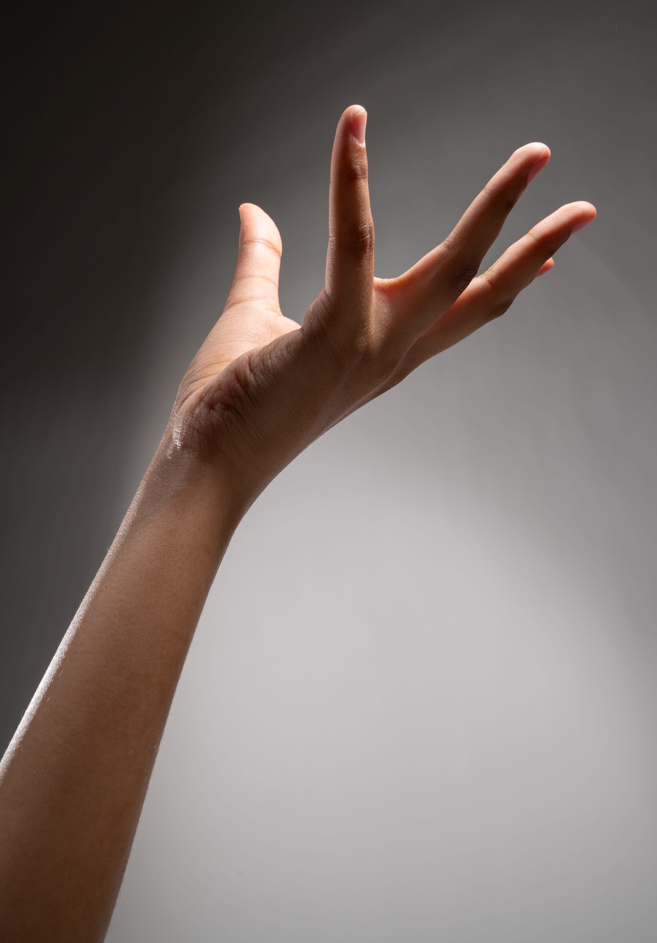 a close up view of the back of an open hand reaching up against a gray background, illuminated by a single source of light.