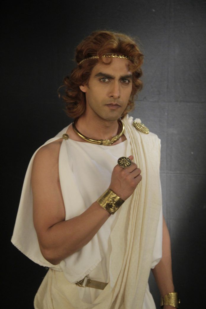 a man dressed in a white toga and gold jewelry, including a headband, necklace, ring, and bracelets. he is posing for a portrait, likely depicting a character from ancient greece.