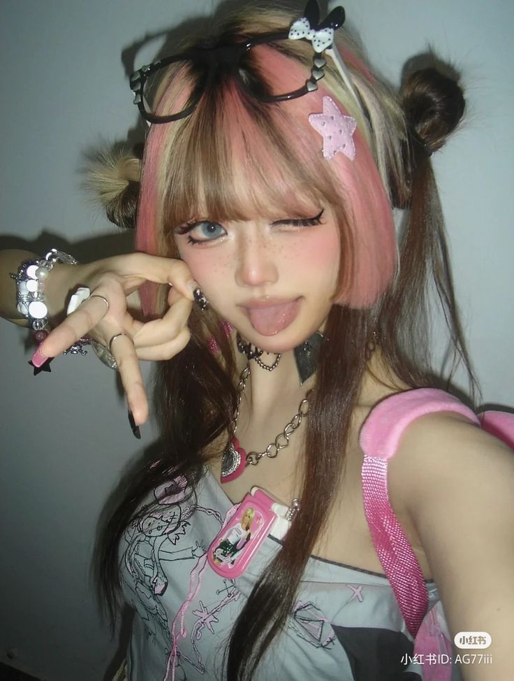 a woman with pink, blonde, and brown hair takes a selfie in a mirror. she is wearing pink and black hair clips, glasses on top of her head, a pink backpack strap, and multiple pieces of jewelry in silver. she is making a playful face at the camera with her tongue out.