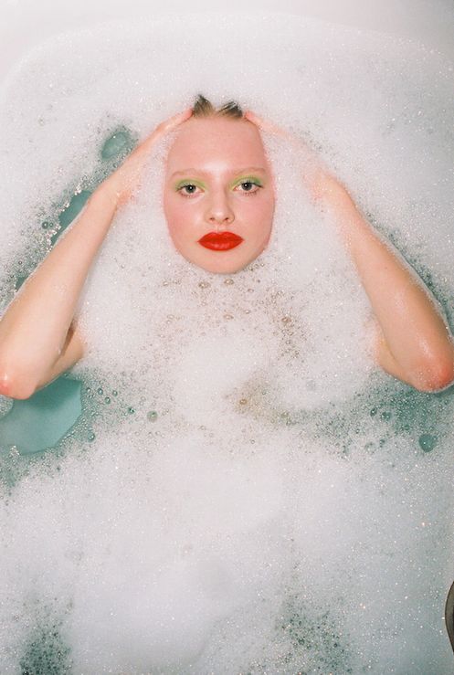 a woman with red lipstick and green eyeshadow is in a bathtub full of bubbles. she has her arms up above her head as if she is relaxing in the bath.  the photo is shot from an overhead perspective.