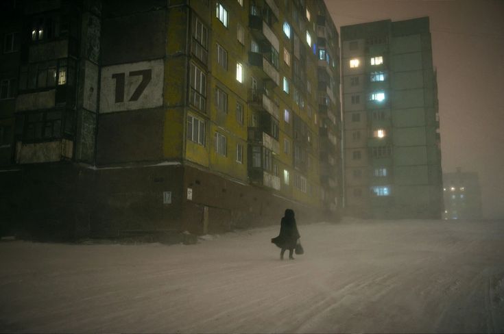 the image depicts a woman in a dark coat walking alone down a snowcovered street at night in what appears to be a russian city.  the architecture is characteristic of sovietera apartment blocks, showing aged, multistory buildings with dimly lit windows.  the scene is atmospheric and moody, with heavy snowfall or a blizzard creating a hazy, almost ethereal effect.  the overall impression is one of isolation, loneliness, and a cold, bleak urban environment.  the number "17" is clearly visible on the side of one of the buildings. the scene is quiet and peaceful, conveying a sense of solitude and stillness.
