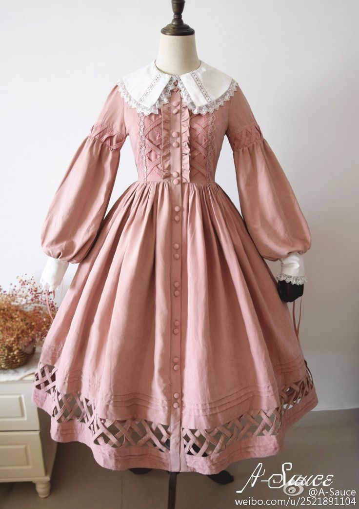 a fulllength photo of a pink lolita fashion dress displayed on a mannequin. the dress features a full skirt, long sleeves with puff shoulders, a lace collar, and an embroidered bodice. the skirt has a lattice pattern around the hem. the dress is a pale pink color with white accents. the background is a white wall.