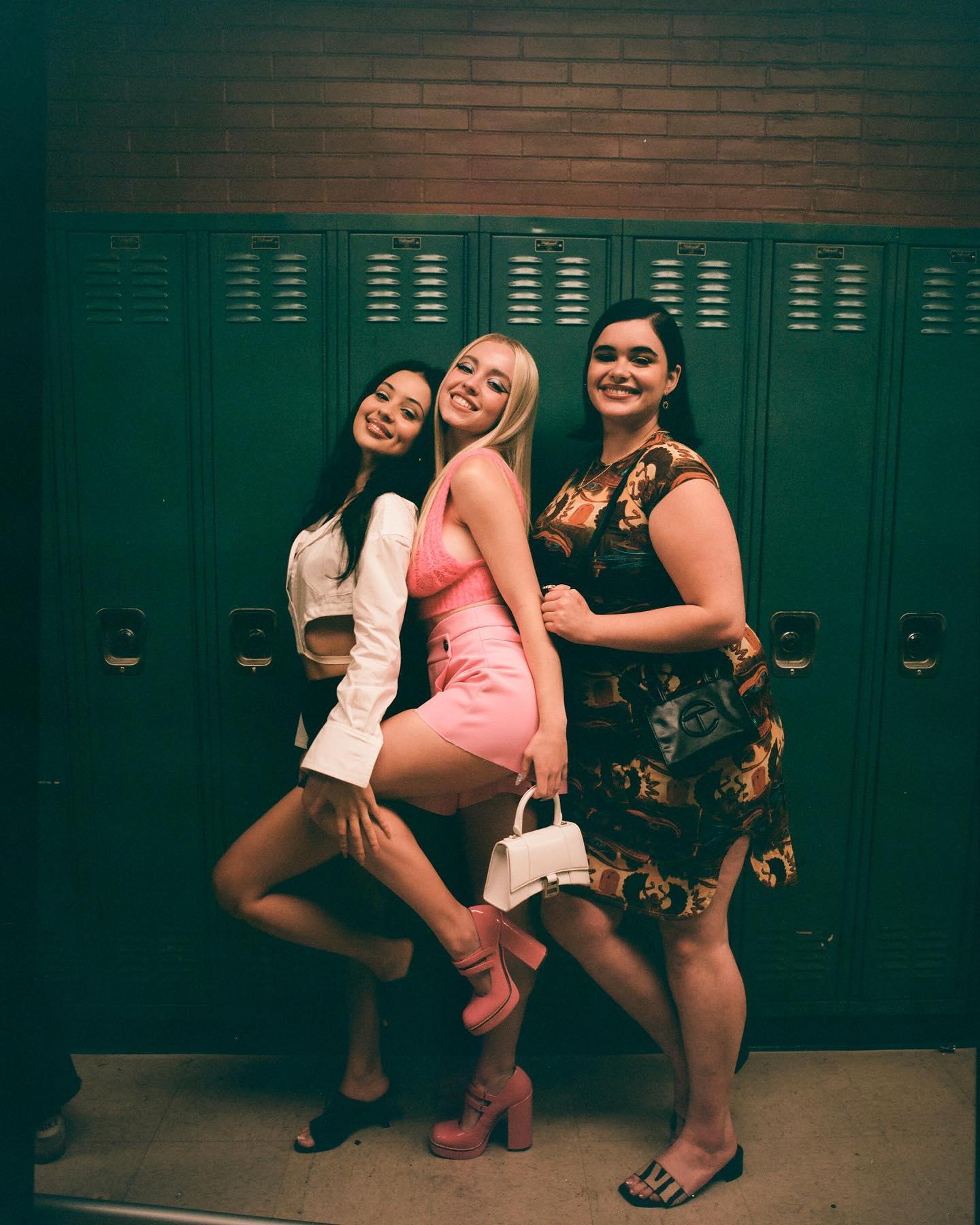 the image shows three teenage girls standing closely together in a high school hallway in front of green lockers.  they are stylishly dressed; one in a pink short set, another in a white shirt and black bottoms, and a third in a patterned dress.  they're all smiling and appear to be friends. the photograph has a vintage filmlike quality, with warm tones and a slightly grainy texture.  the overall vibe is one of casual friendship and youthful style.