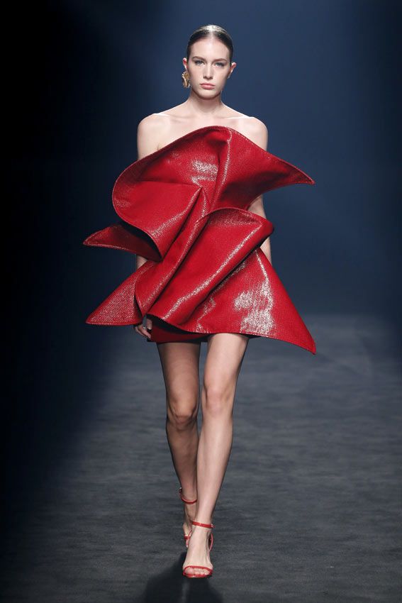 a fashion model walks down the runway in a striking red dress with a unique, sculptural design. her outfit is accentuated by simple red sandals, showcasing the intricate details of the dress. the model's pose and confident stride add to the overall elegance and glamour of the presentation.