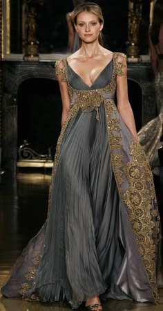 a woman in a stunning gray evening gown with intricate gold lace detailing stands on a fashion runway. the dress is flowy and elegant, with a sweetheart neckline and a fitted bodice. the model's hair is styled in a sleek updo, and her makeup is natural and chic.  she poses confidently, exuding glamour and sophistication. the backdrop is a blurred studio setting, highlighting the beauty of the dress and the model. the overall image portrays a highfashion aesthetic and captures the essence of elegance and femininity.