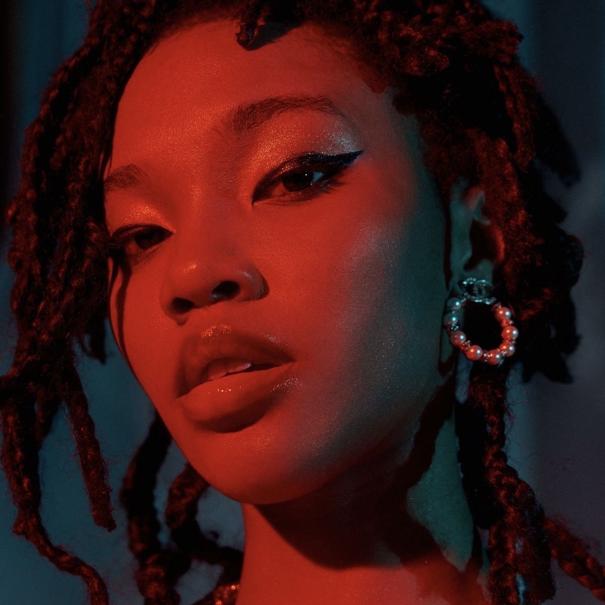 a closeup portrait of a woman with red hair and red skin. she is wearing earrings and makeup, and her face is lit in a red and blue light. her skin and lips are highlighted in red and her eyes are dark and smoky, creating a glamorous and edgy effect.