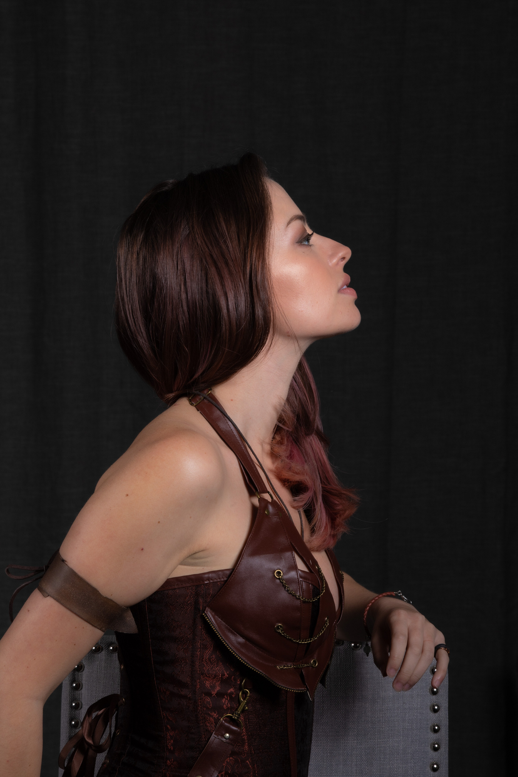 a woman with long reddishbrown hair is shown in profile view against a black backdrop. she is wearing a brown leather steampunkinspired corset top with intricate detailing and a matching arm band. her makeup is bold and she gazes confidently offcamera. the lighting is soft and highlights her features, while the overall mood is sensual and powerful. she rests her arm on a gray chair, partially visible on the right edge of the frame.