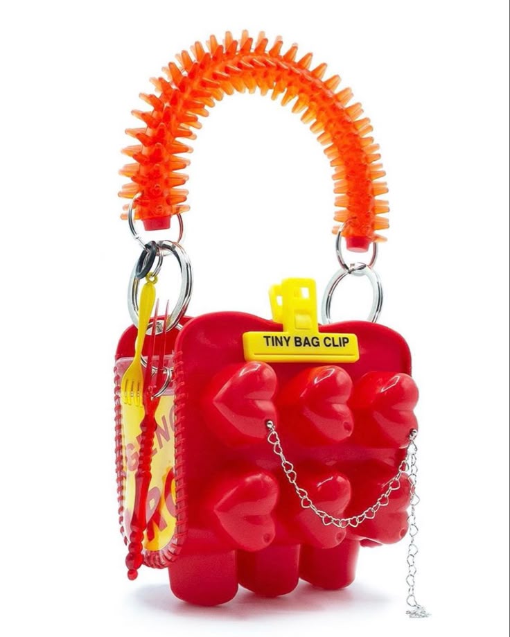 a closeup studio shot of a novelty handbag on a white background. the body of the bag is red and made of plastic, featuring rows of raised hearts. a yellow plastic bag clip with the words "tiny bag clip" is attached to the top. the handle is orange and made of textured plastic, reminiscent of a spiky massage tool. a silver chain drapes down the side of the bag. a yellow plastic fork and spoon set is visible inside, suggesting it may be intended for carrying small items. the handbag has a playful, unconventional design.