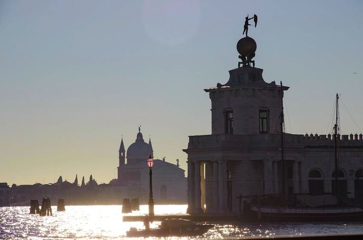 a silhouette of a venetian cityscape at sunset. the main focus is on a stone building with a statue on top, situated on the water. in the background, a church is visible, and the setting sun casts a golden glow on the water. the overall mood is peaceful and serene.