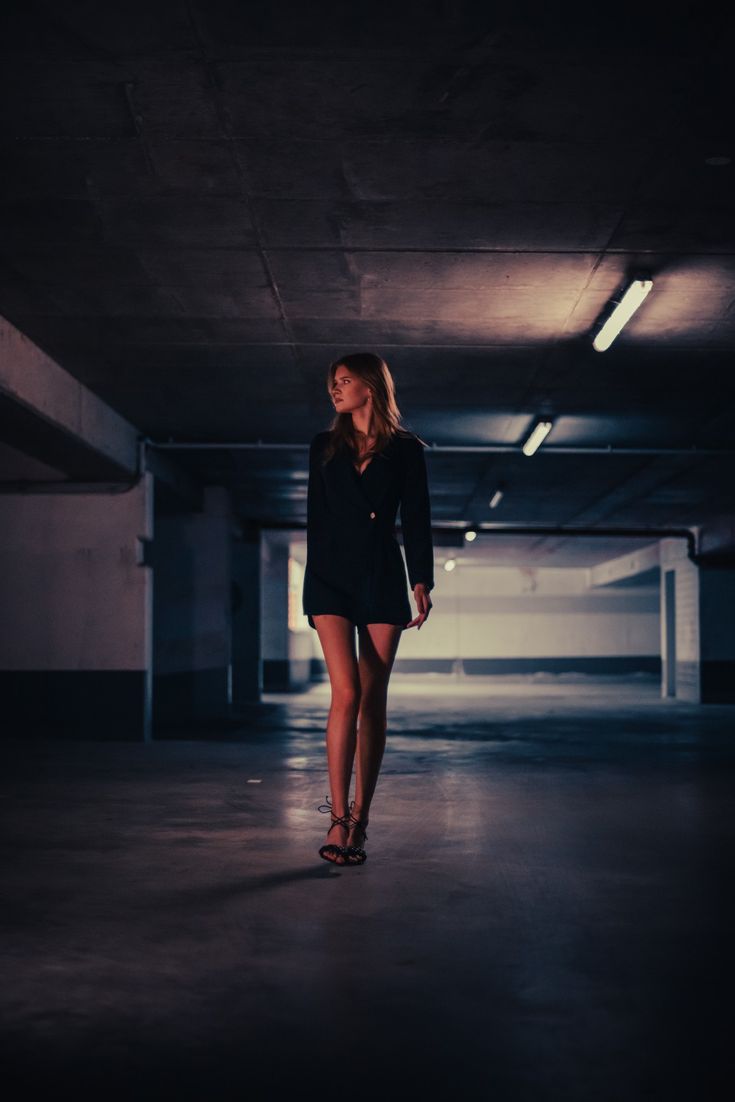 a young woman is walking in a parking garage. she is wearing a black blazer dress and heels. the concrete is dark and there is a lot of shadow.  she is looking off to the side.