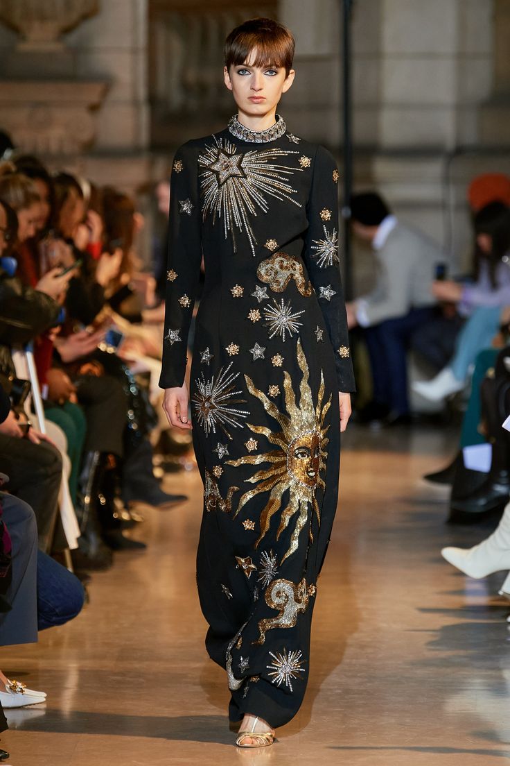 a model walks the runway in a stunning black long gown embellished with celestial designs. the dress features intricate embroidery of stars, a sun, and other celestial elements in gold and silver.  the model has short hair and is wearing minimal jewelry. the background shows a blurred view of other people watching the fashion show. the overall mood is elegant and sophisticated, showcasing the luxury of high fashion.
