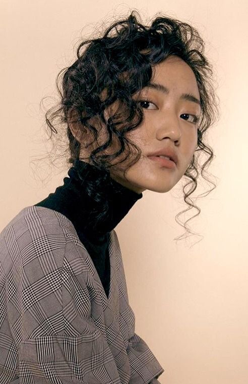 a young woman with curly hair and freckles poses for a portrait. she wears a black turtleneck and a gray plaid dress. the background is a light beige. she is lit with soft lighting, creating a natural look.  the perspective is at eye level.