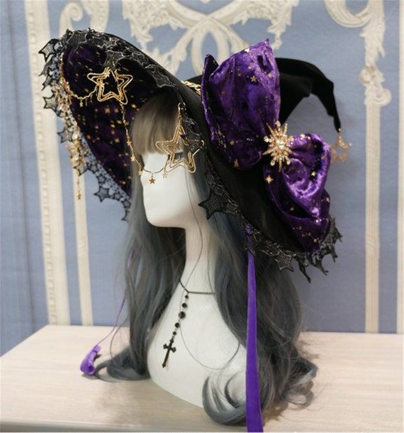 a closeup shot showcases a decorative witch hat atop a mannequin head. the hat is primarily black, embellished with purple accents in a velvety fabric featuring gold star patterns. a large, plush bow adorns one side, secured with a starshaped ornament. the hat's brim is lined with intricate black lace details, further decorated with dangling gold star charms. a gold star headpiece is worn by the mannequin. the mannequin has grey hair and wears a necklace with a cross pendant. the background is a patterned blue and white backdrop.