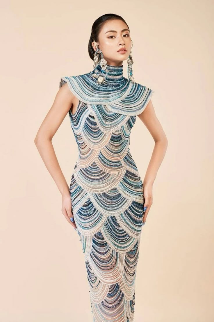 a woman is posing in a floorlength gown with teal, cream, and blue beadwork in a scalloped design. the dress features a high neckline and sleeveless design. she is wearing large, elaborate earrings and her hair is pulled back in a bun. she has a neutral expression and her hands are at her sides. the background is a light brown color.