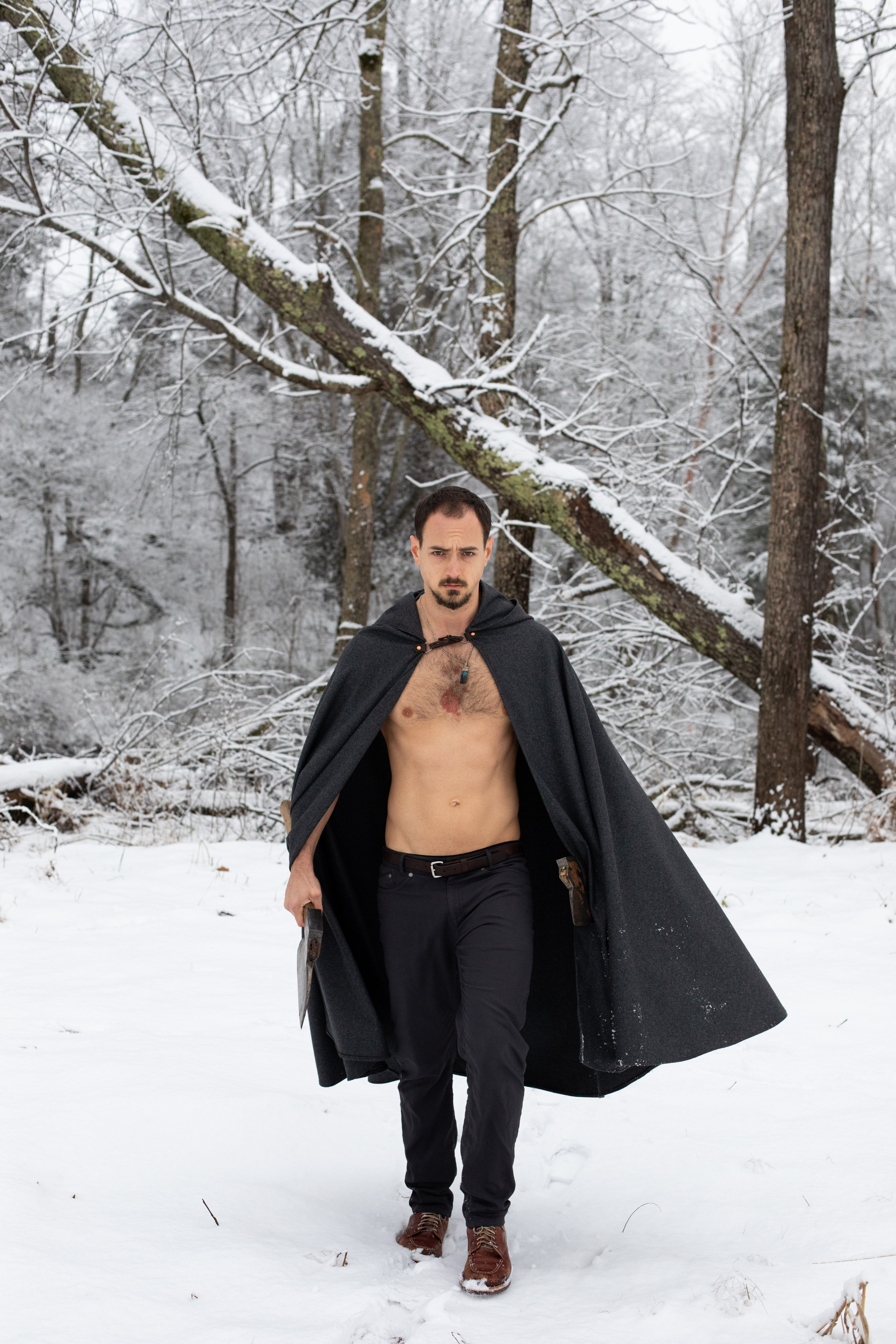 a man with a bare chest, wearing a black cape and carrying an axe, walks through a snowy forest. he has a determined look on his face. the scene is set in winter, with snow covering the ground and trees.