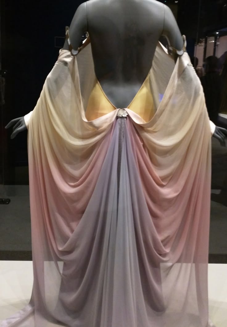 a back view of a mannequin wearing a long, flowing gown with an ombré effect transitioning from light yellow at the top to pink and then lavender towards the bottom. the dress is made of chiffon fabric with draped detailing. the mannequin is sleeveless and backless, and a gold fabric accent is visible behind the draped portion of the dress. the overall impression is of a highfashion garment displayed in an exhibition setting.