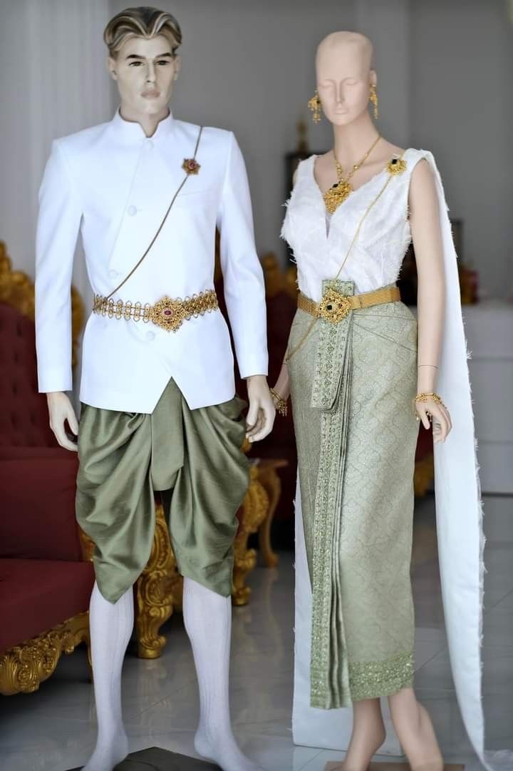 a thai bride and groom dressed in traditional wedding attire. the groom is wearing a white longsleeved jacket with gold trim and a gold belt over green pants. the bride is wearing a white blouse with gold trim and a green long skirt. they are both smiling and looking at the camera. the background is a white wall with gold trim and a gold throne.