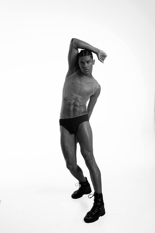 a black and white studio shot of a man in a fashionable pose. he is wearing a mesh top, black swimwear, and black boots. his right arm is raised above his head.