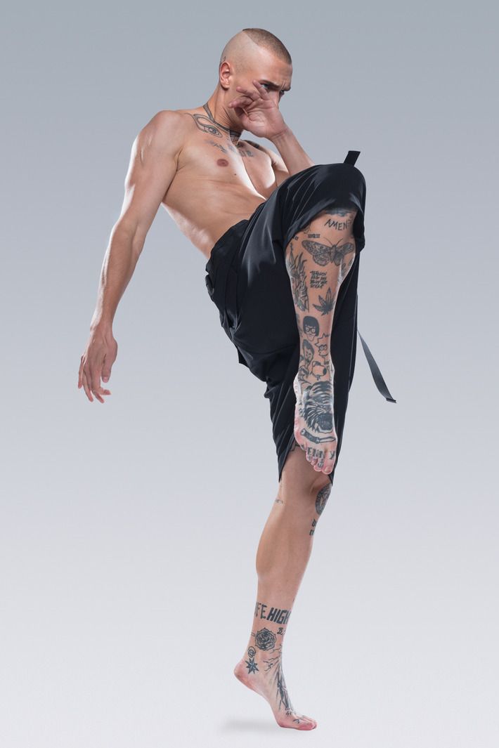 a young, tattooed male model is posing in a studio. he is wearing black shorts, and has his right leg lifted, foot pointing towards the camera. he is looking down and has one hand on his face.  the image has a dark and moody feel.