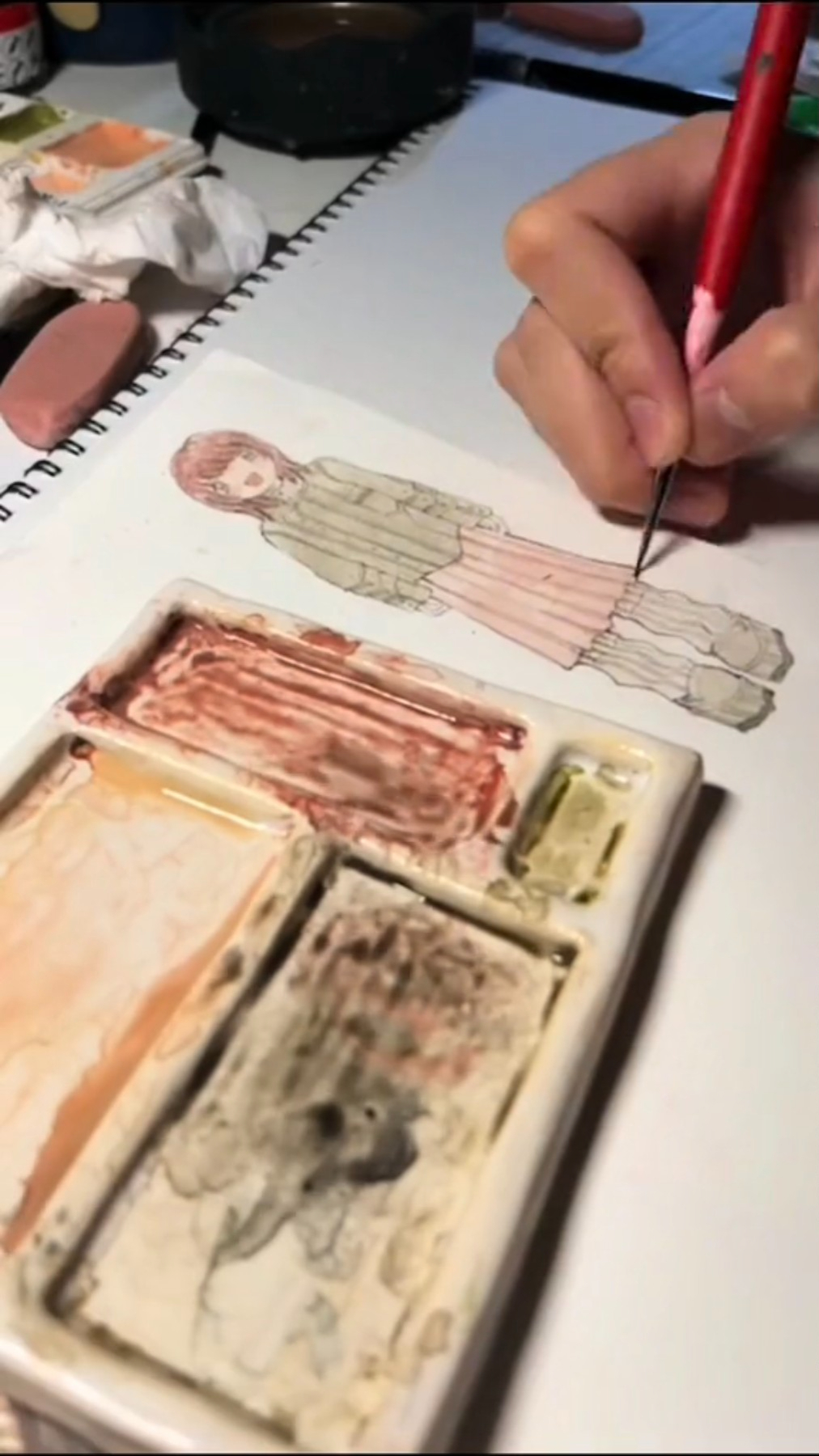 a person is drawing a girl wearing a dress and shoes. they are using a red watercolor brush and a water color palette that is filled with watercolors. the person is holding the brush with their right hand. they are seated at a table and their other hand is out of frame. they are using a white piece of paper for their art. the drawing of the girl is incomplete.
