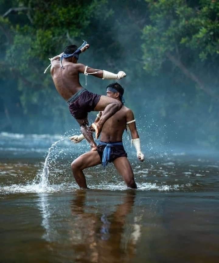 two shirtless men are engaged in a muay thai fight in a river. the man on top has his leg up and is kicking the man on the bottom. the man on the bottom is blocking the kick with his arm and is getting ready to counter attack. both men are covered in sweat and are in a serious stance.
